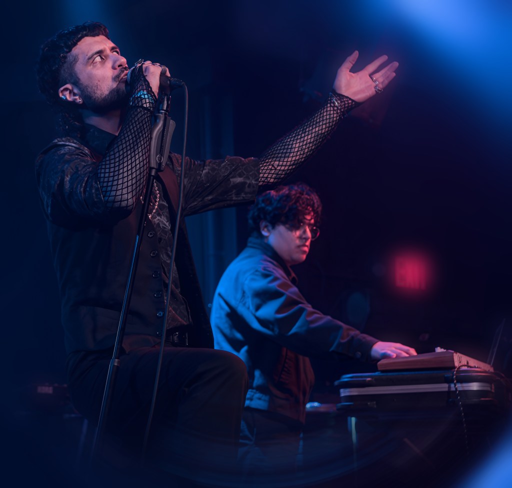 A male singer passionately performing on stage with a microphone, while a male musician plays electronic instruments in the background, illuminated by stage lights.