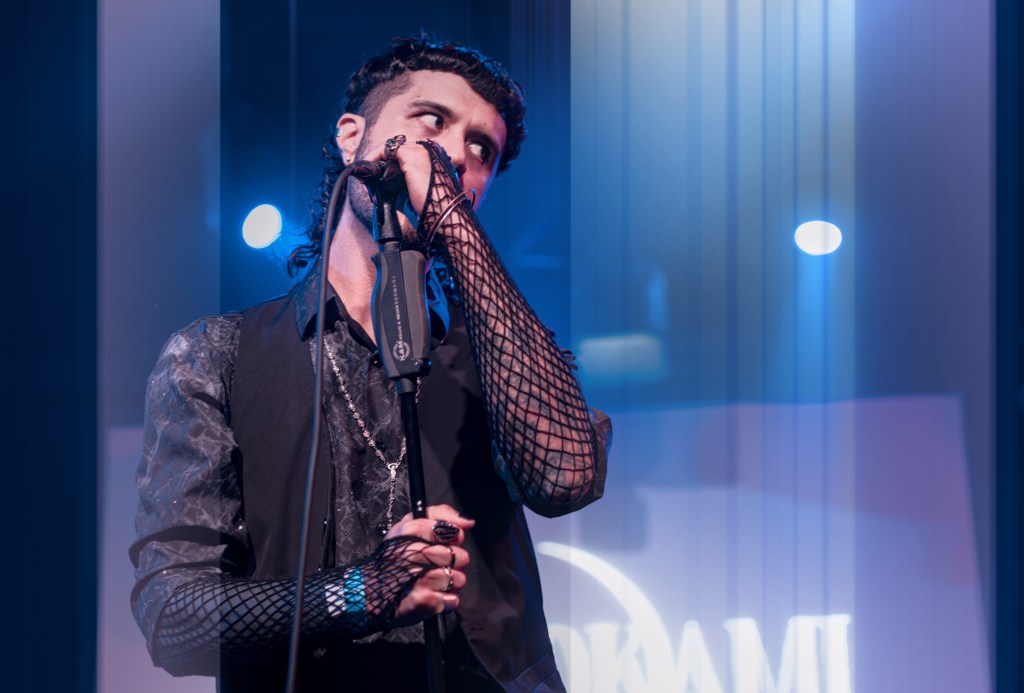 A musician performing on stage, holding a microphone with a dramatic expression. They are dressed in a stylish outfit featuring a fishnet top and a tailored vest, surrounded by blue stage lights.