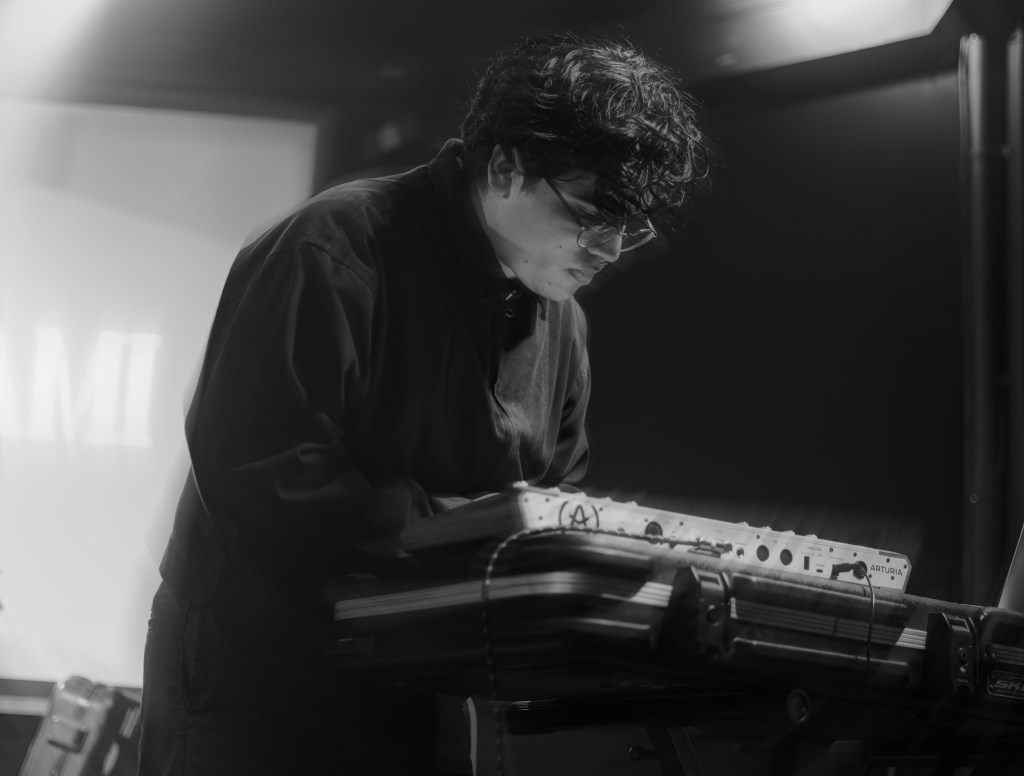 A musician performing with a keyboard on stage, captured in black and white. They have curly hair, glasses, and are focused on their performance.