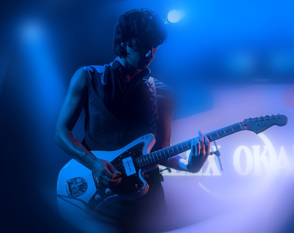 A musician playing an electric guitar on stage under blue lighting.