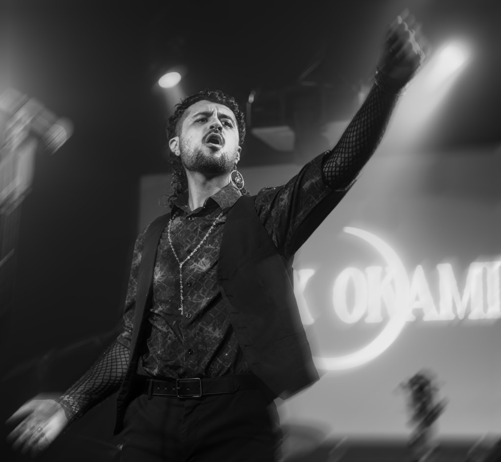 A male performer passionately singing on stage, wearing a stylish black outfit with intricate patterns. He has curly hair and is adorned with accessories, expressing intense emotion during a live performance.