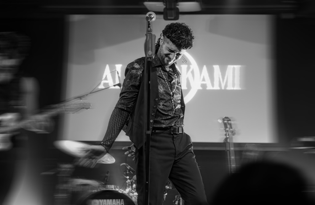 A black and white image of a male singer performing on stage, wearing a patterned shirt and holding a microphone stand. The background features a logo that reads 'AKAMI'.