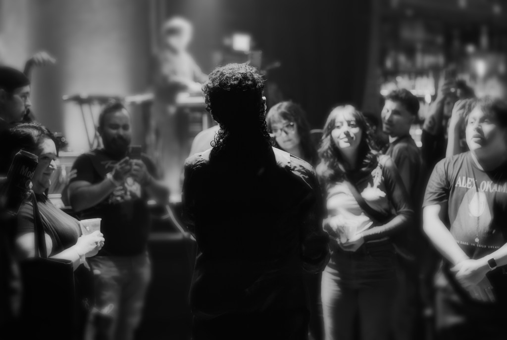 A black and white image of a crowd at a music event, focusing on a performer with their back to the camera as they engage with an audience. The attendees, including men and women, are visibly enjoying the performance and interacting with each other.