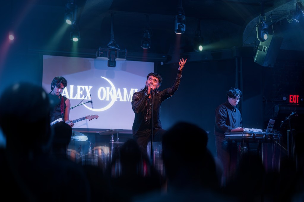 A live performance with a male singer passionately gesturing on stage, accompanied by a guitarist and a keyboardist, under colorful stage lights with a backdrop displaying the name 'ALEX OKAMO'.