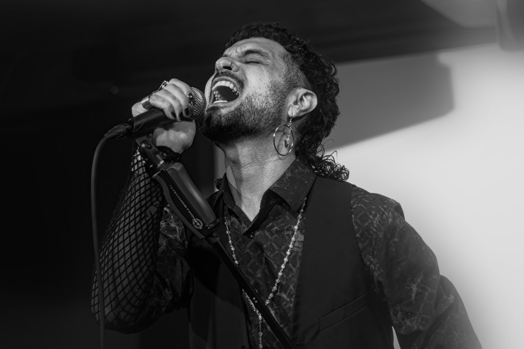 A passionate male vocalist singing into a microphone, with an intense expression, wearing a black vest and a patterned shirt. The image is in black and white.