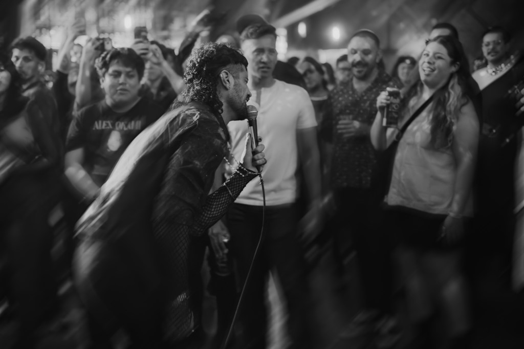 A performer singing passionately into a microphone in front of an engaged crowd at a lively concert, captured in black and white.