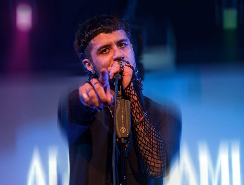 A performer on stage pointing towards the audience while holding a microphone, dressed in a black outfit with fishnet sleeves, illuminated by colorful stage lights.