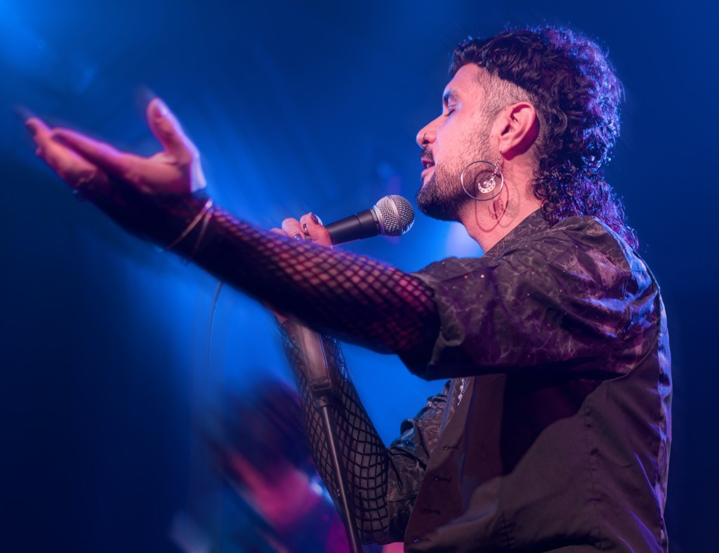 A performer passionately singing into a microphone on stage, with a hand extended and wearing a stylish outfit including fishnet sleeves.