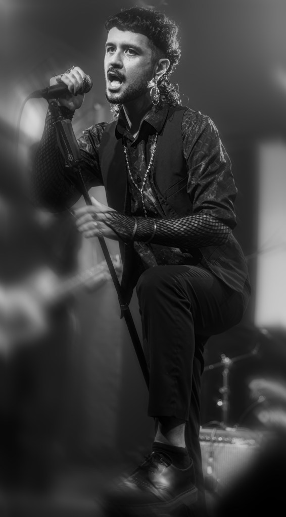 A singer performing on stage, passionately holding a microphone and dressed in stylish attire with accessories, exuding energy and emotion during a live music event.