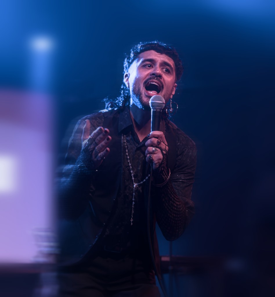 A performer passionately singing into a microphone on stage, wearing a black outfit with mesh sleeves and accessories, set against a blue lighting backdrop.