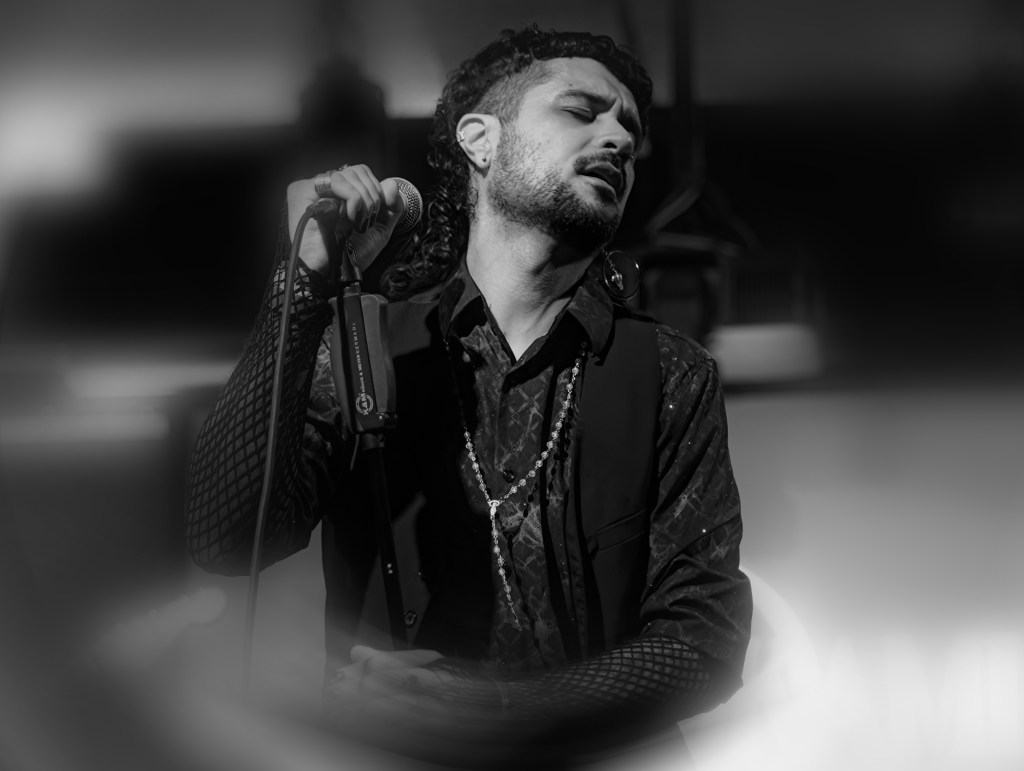 Singer performing passionately on stage, holding a microphone, dressed in a stylish black outfit with intricate details.