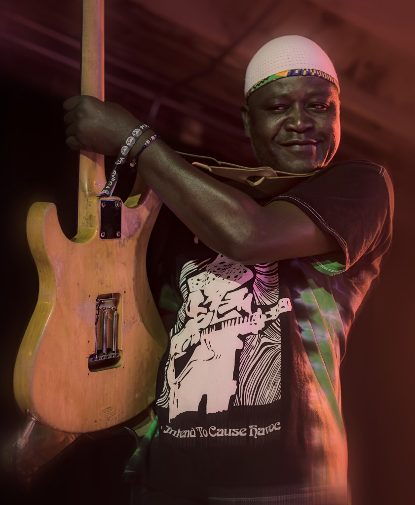 A musician holding an electric guitar, smiling while wearing a patterned t-shirt and a white cap.