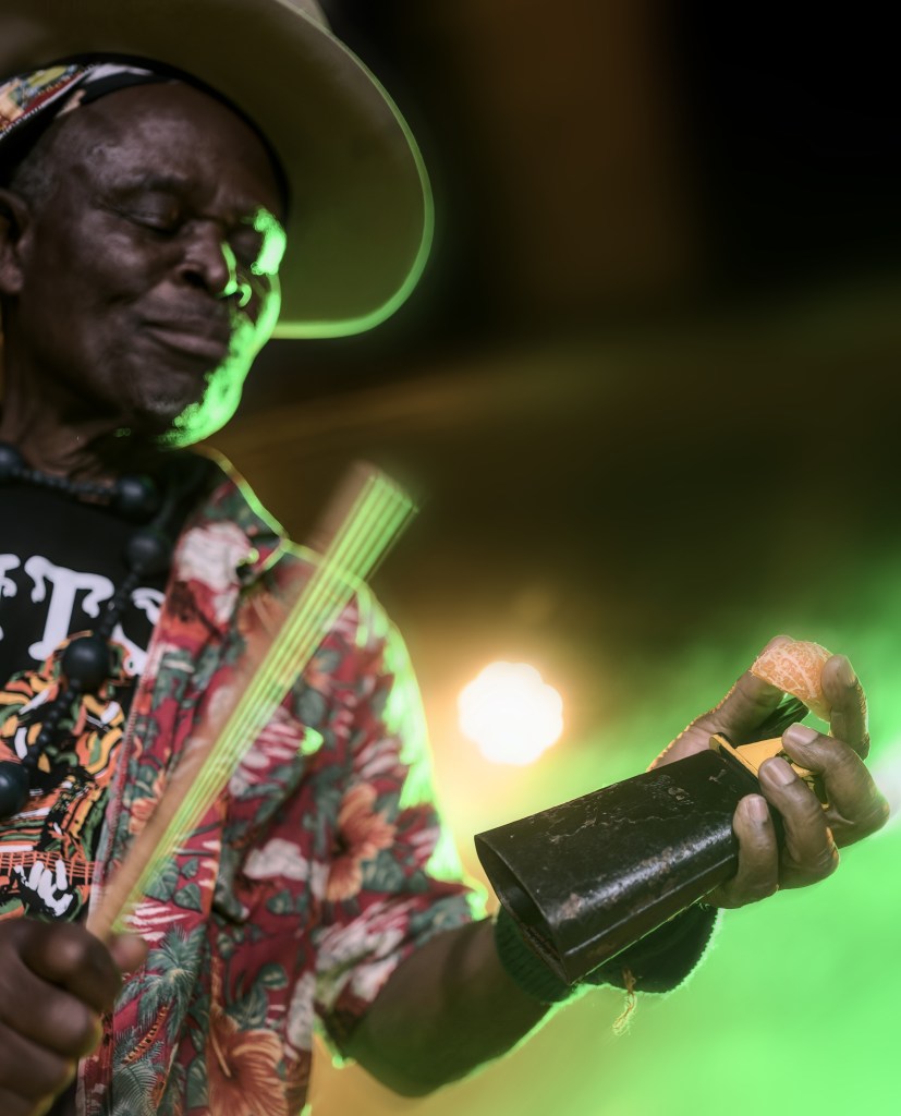 An elderly musician wearing a colorful shirt and a hat, holding a musical instrument in one hand and a piece of fruit in the other, performing on stage with green lighting in the background.