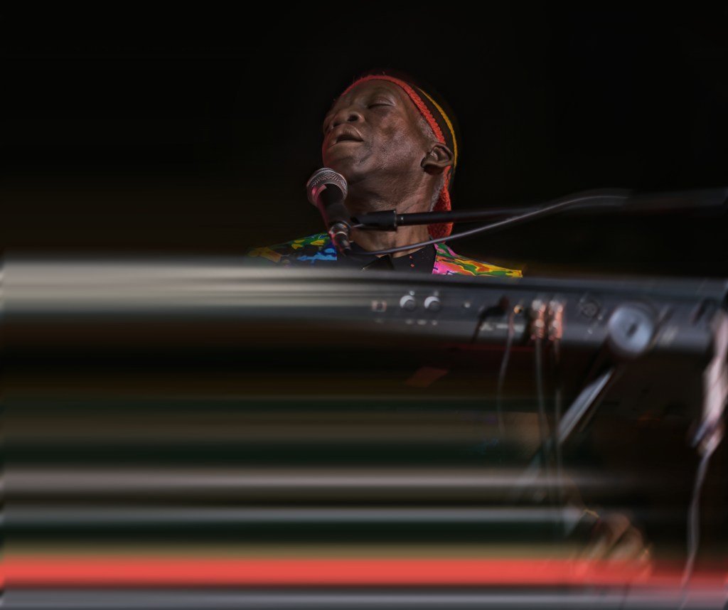 A musician passionately performing at a keyboard, wearing a colorful, patterned shirt and a headband, with eyes closed and microphone nearby.