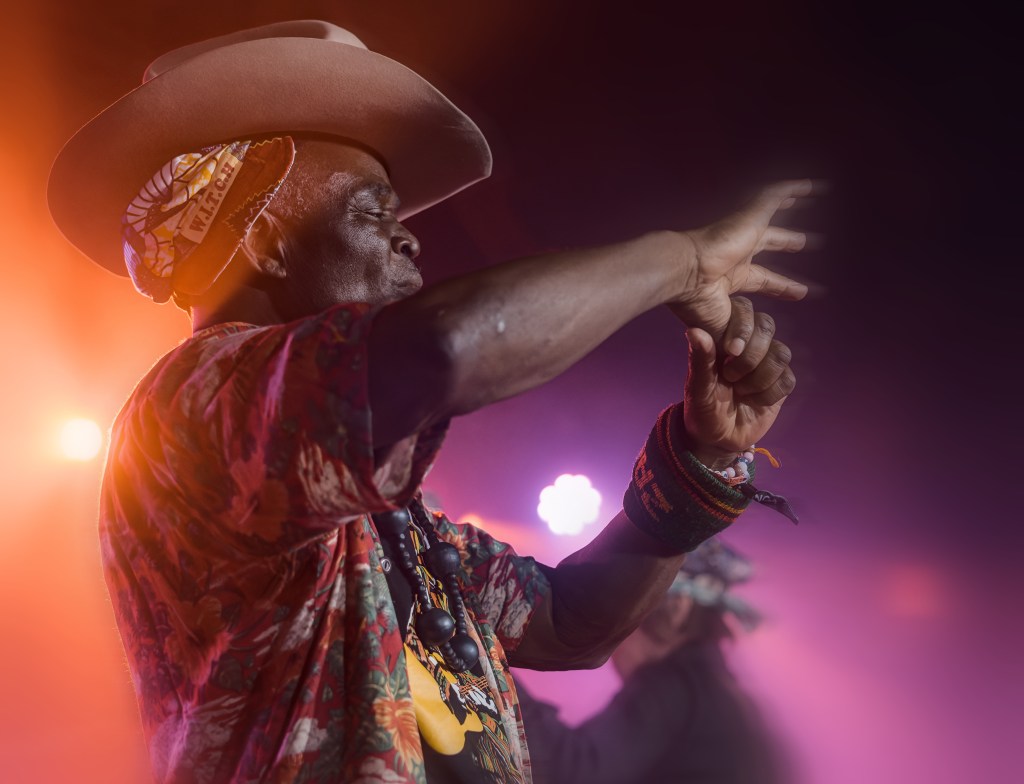 A performer in a colorful floral shirt and a wide-brimmed hat gestures expressively while on stage, with colorful lights in the background.