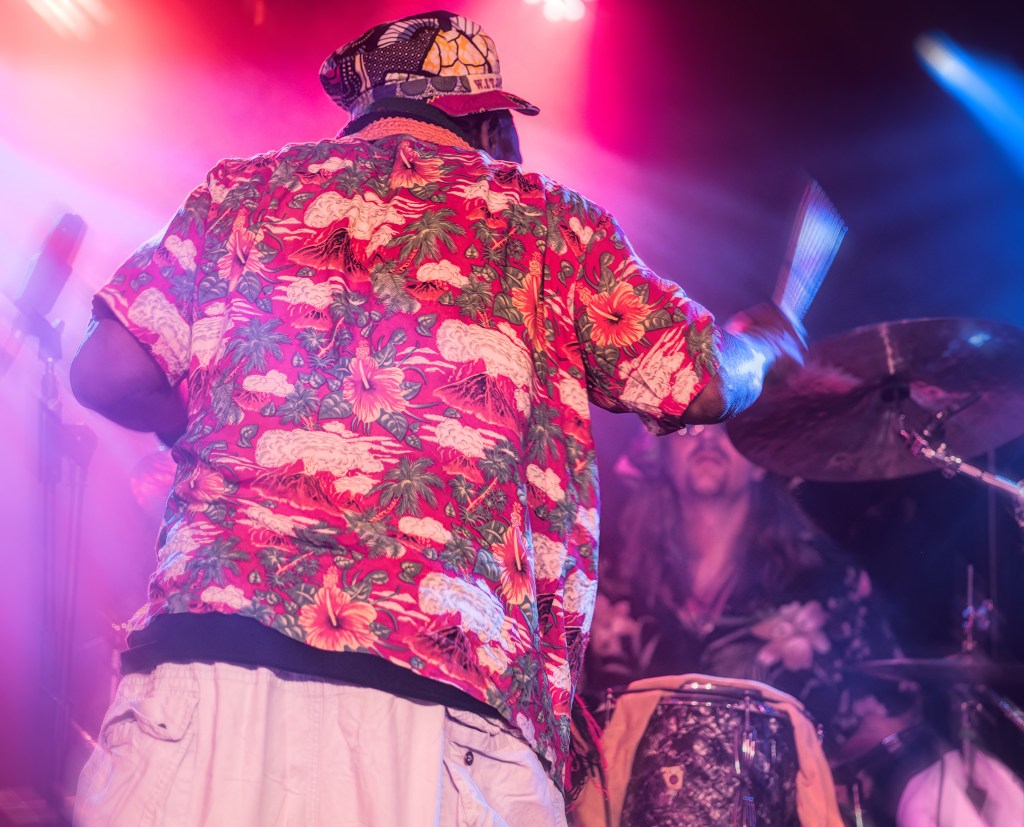 A drummer wearing a colorful floral shirt and hat performs on stage, with vibrant lighting in the background.