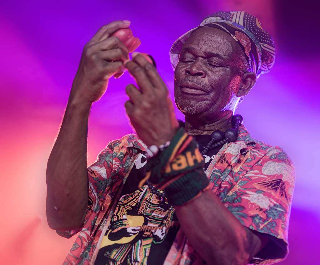 An elderly musician performs on stage, focused on his music, with colorful stage lighting illuminating him. He wears a vibrant floral shirt, a patterned hat, and has beaded accessories.