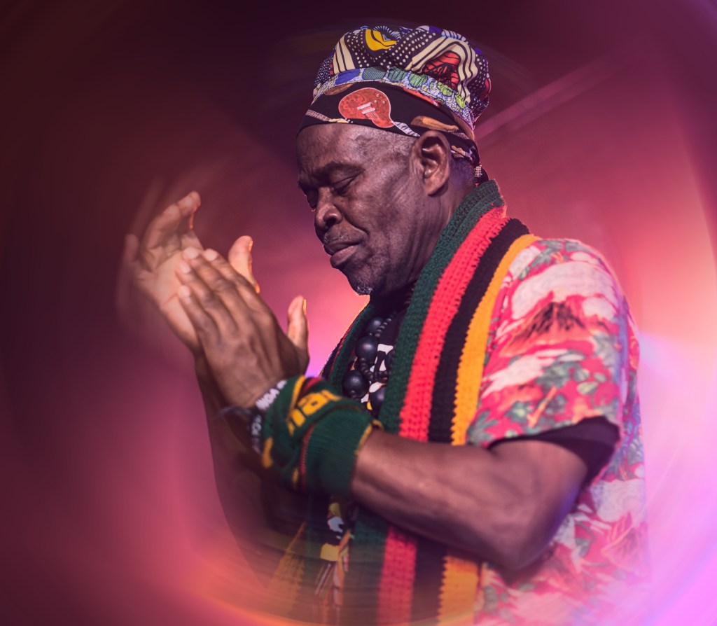 A man wearing a colorful patterned shirt and headscarf, performing passionately on stage with his eyes closed and hands raised, surrounded by vibrant lighting.