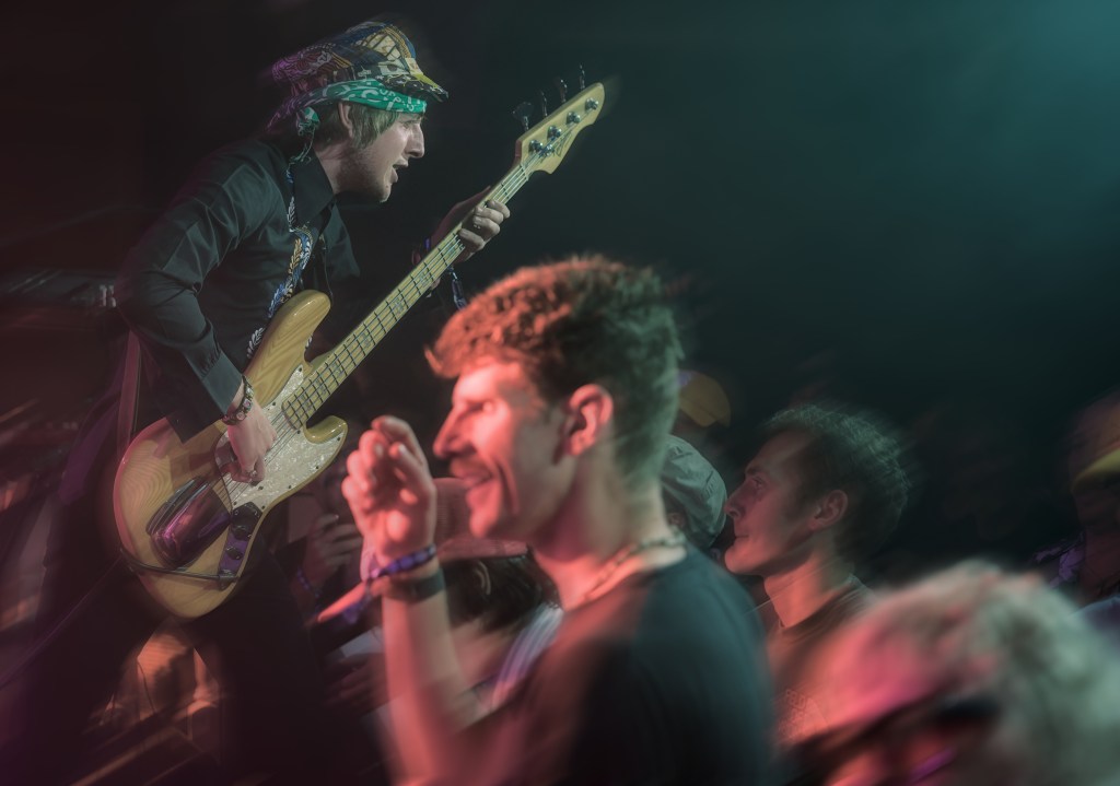 A musician passionately playing a bass guitar on stage, surrounded by an enthusiastic crowd, with some audience members visibly engaged and dancing.