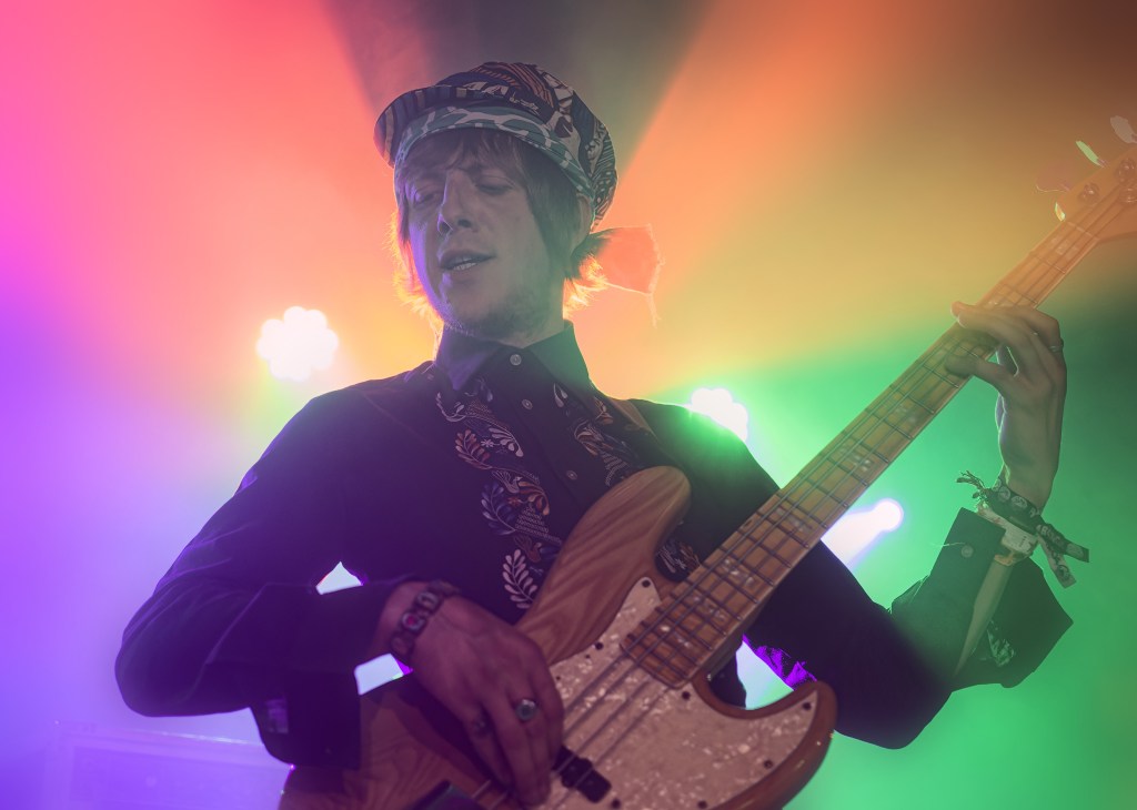 A musician playing a bass guitar under colorful stage lights, wearing a patterned shirt and a hat.