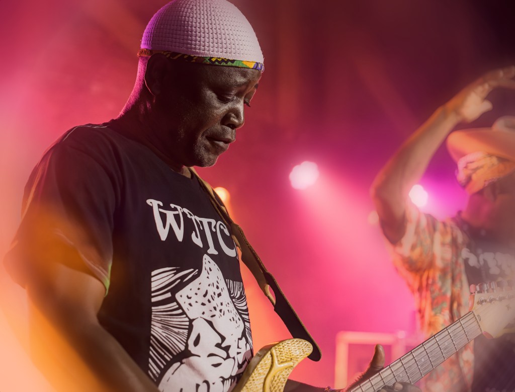 A musician playing an electric guitar on stage, wearing a black T-shirt and a white head wrap, with colorful stage lighting in the background.