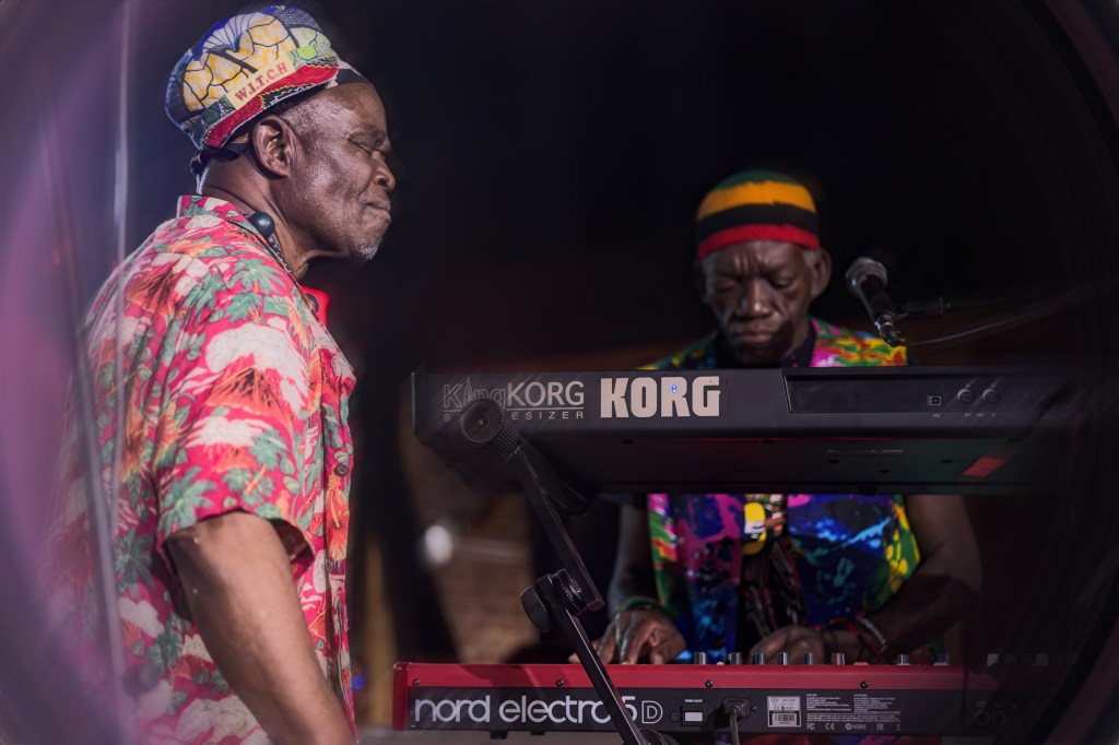 Two male musicians on stage, one playing a keyboard and the other singing, both wearing colorful shirts and hats.