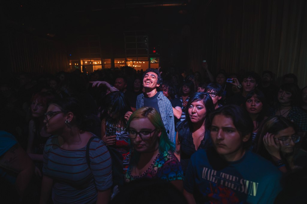 A diverse crowd at a concert, with one person in the foreground smiling and enjoying the performance, while others appear focused or serious.
