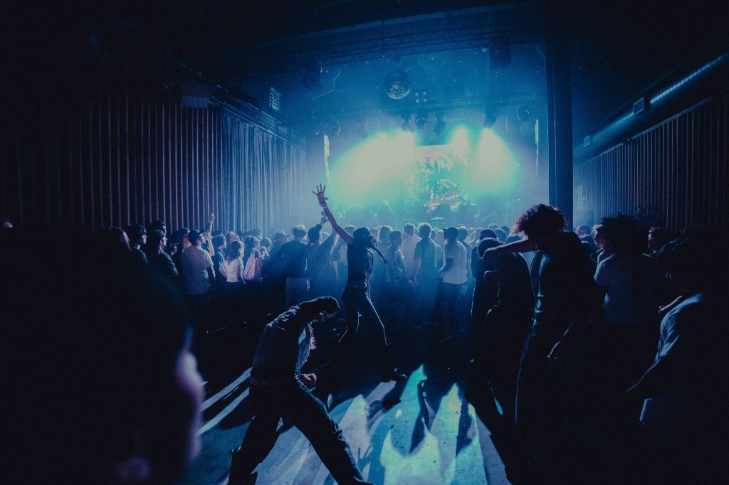 A lively crowd dancing at a concert with colorful stage lighting in a dark venue.