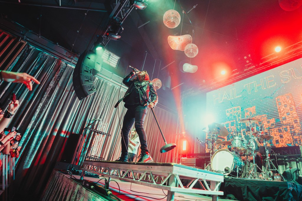 A lead singer performing on stage with a microphone, wearing a black leather jacket, while a drummer is visible in the background. The stage is illuminated with colorful lights, and the crowd is engaged in the performance.