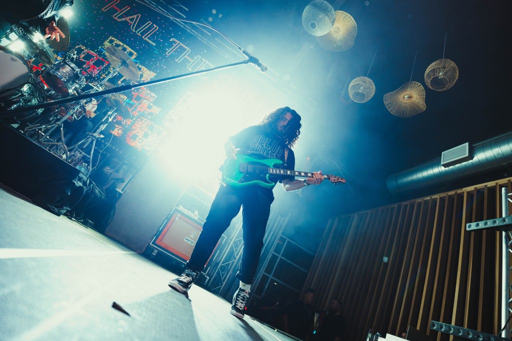 A guitarist performing on stage with bright stage lights and fog. The musician has curly hair and is playing a green electric guitar. Drums and amplifiers are visible in the background.