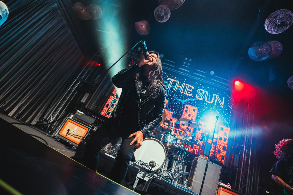 A singer performing on stage with a microphone, wearing a leather jacket, in front of a vibrant backdrop featuring colorful lights and the text 'THE SUN'.