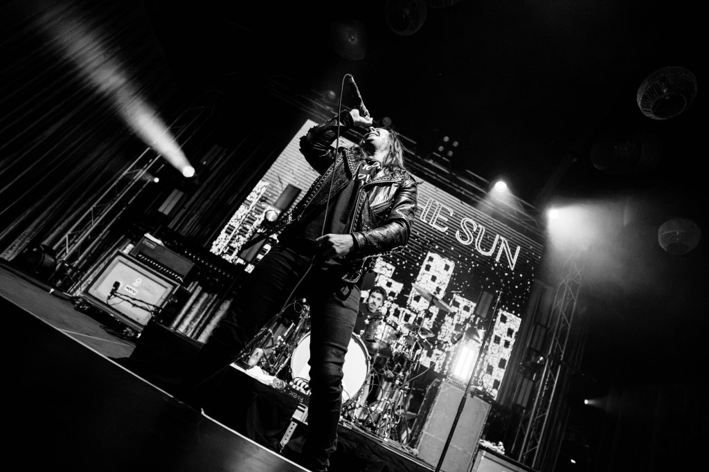 A vocalist performing on stage in a black leather jacket, with dramatic lighting and a band setup in the background.