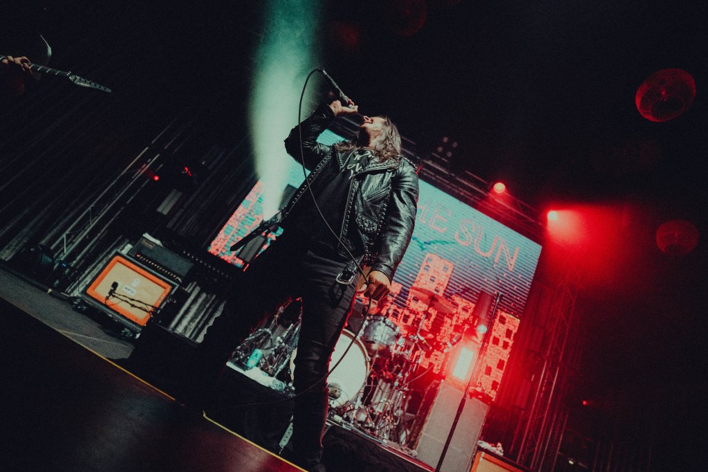 A musician performing on stage with a microphone, wearing a leather jacket, with vibrant red and blue stage lights in the background and a visible drum set.