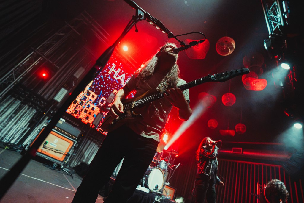 A musician playing electric guitar on stage with a dynamic lighting setup, including red lights and a vibrant background display. Other band members are visible, performing in the background.