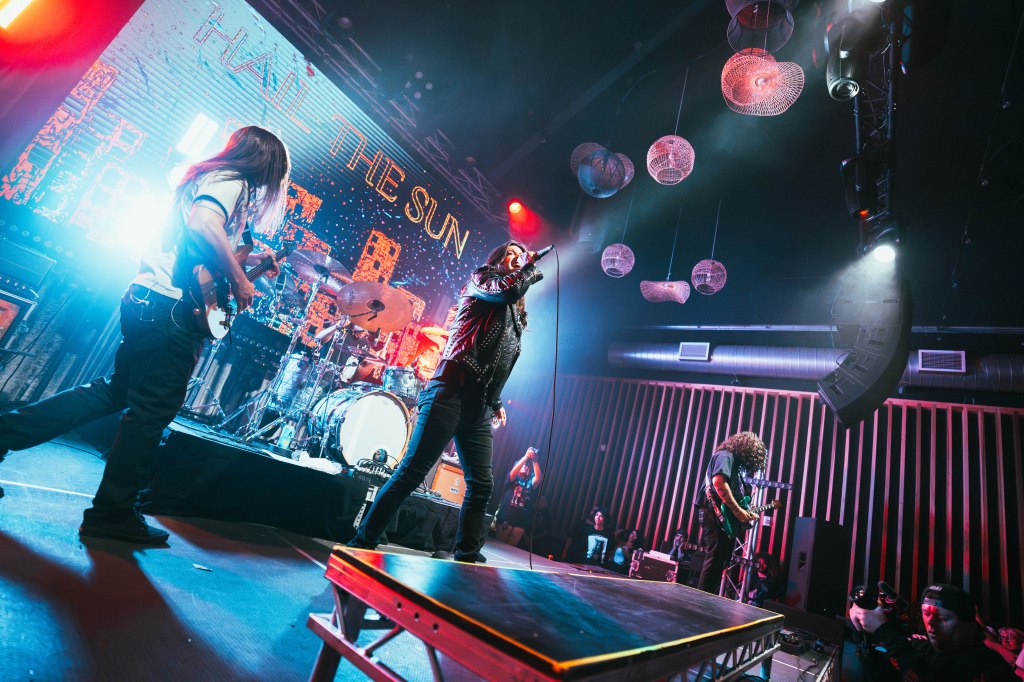 A live music performance featuring a band on stage, with a vocalist in a leather jacket and guitarists playing instruments. The backdrop displays the text 'HAIL THE SUN', illuminated by stage lights, creating an energetic atmosphere.