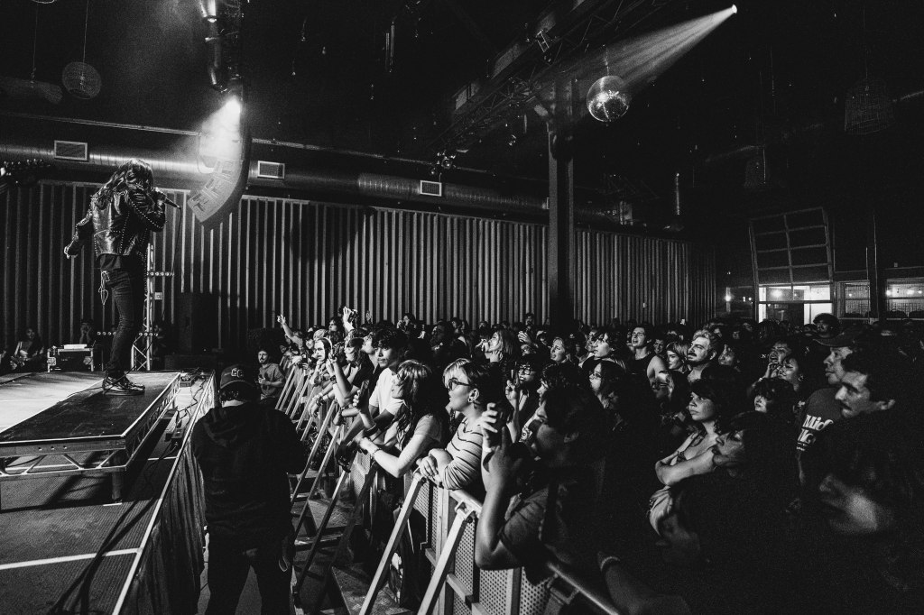 A performer in a leather jacket sings on stage in front of an enthusiastic crowd at a concert, with dramatic lighting and shadows creating an energetic atmosphere.