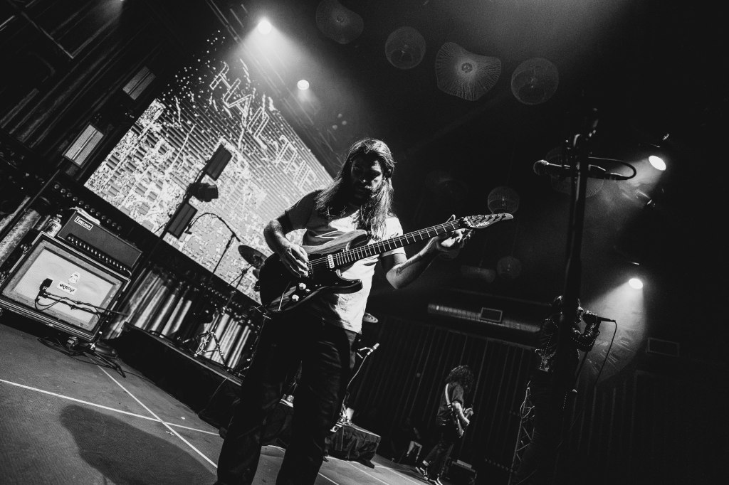 A guitarist performing on stage with an electric guitar, illuminated by stage lights, with a band setup visible in the background and a digital screen displaying graphics.