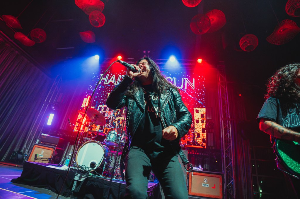 Lead vocalist passionately performing on stage with a microphone, wearing a leather jacket, with colorful stage lights in the background.