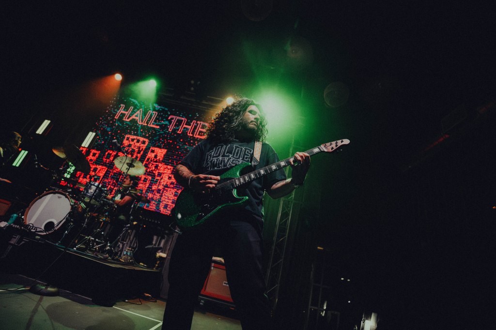 A guitarist performing on stage with a green guitar, illuminated by colorful stage lights and a digital backdrop displaying the words 'HAIL THE'.