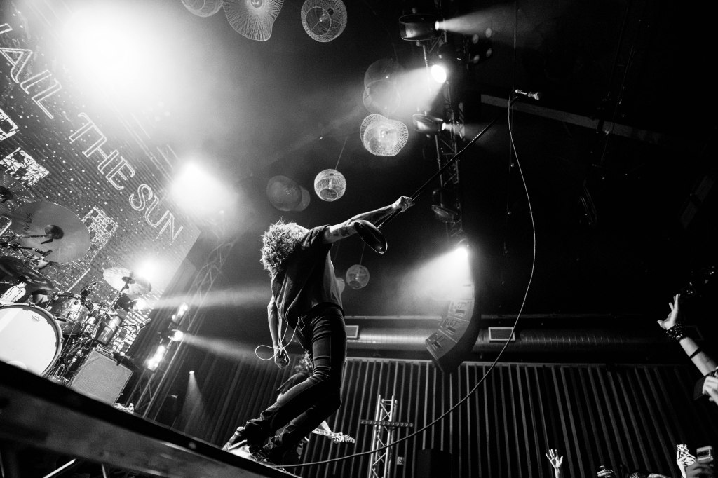 A dynamic black and white image of a musician performing on stage, holding a microphone with one hand while appearing to leap into the air. The background features stage lights and a banner that reads 'HAIL THE SUN.'