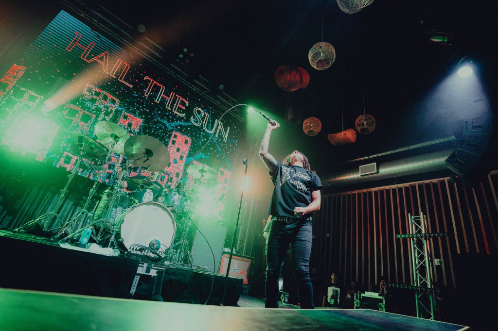 A musician performs on stage in front of a vibrant backdrop with the words 'HAIL THE SUN.' The performer holds a microphone high and is surrounded by colorful lighting, with a drummer in the background.