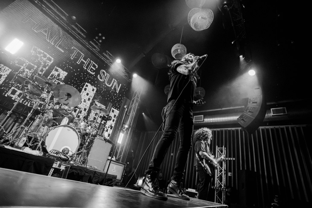 A live music performance featuring a vocalist on stage with a microphone, expressing energy. In the background, a drummer is visible along with a guitarist. The backdrop displays the band name 'Hail the Sun' with vibrant lighting creating an energetic atmosphere.