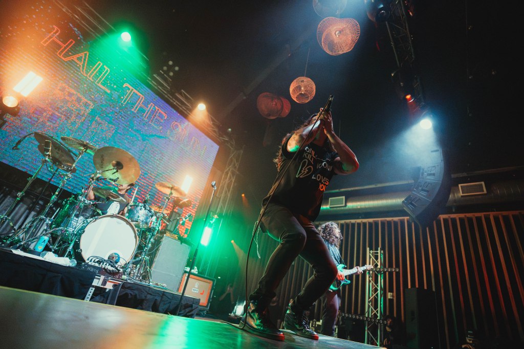 A live music performance featuring a band on stage with colorful lighting. The lead singer is holding a microphone, passionately engaging with the audience, while a drummer and guitarist are visible in the background.