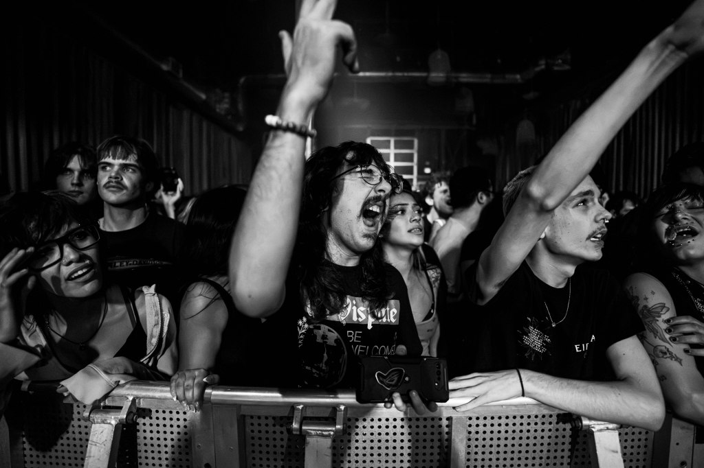 A crowd of enthusiastic concertgoers at a live music event, with diverse expressions of excitement and energy, holding onto a barrier in a dimly lit venue.
