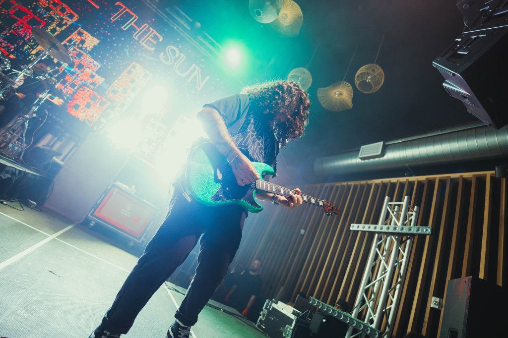 A guitarist performing on stage with a colorful electric guitar, surrounded by dramatic lighting and a band setup in the background.