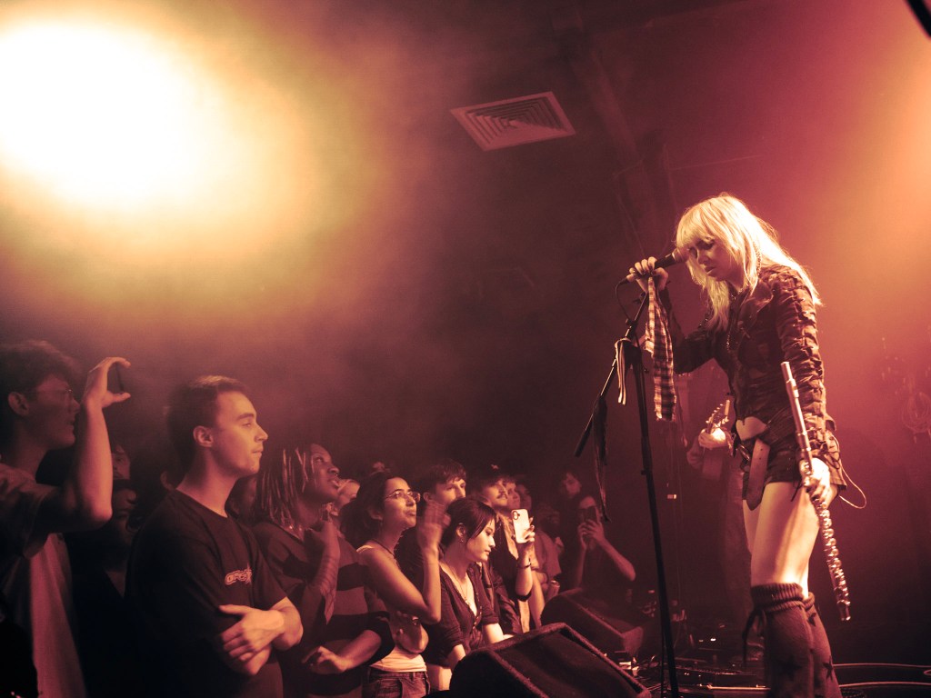 A female performer with long blonde hair sings into a microphone on stage, surrounded by a captivated audience. The lighting is dim with an orange hue, creating a vibrant atmosphere. Fans are seen cheering and taking photos.