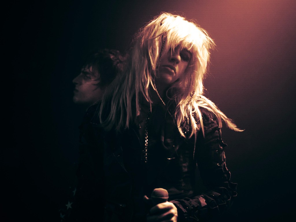 A singer with long, messy hair is holding a microphone while performing on stage, with a shadowy figure in the background.