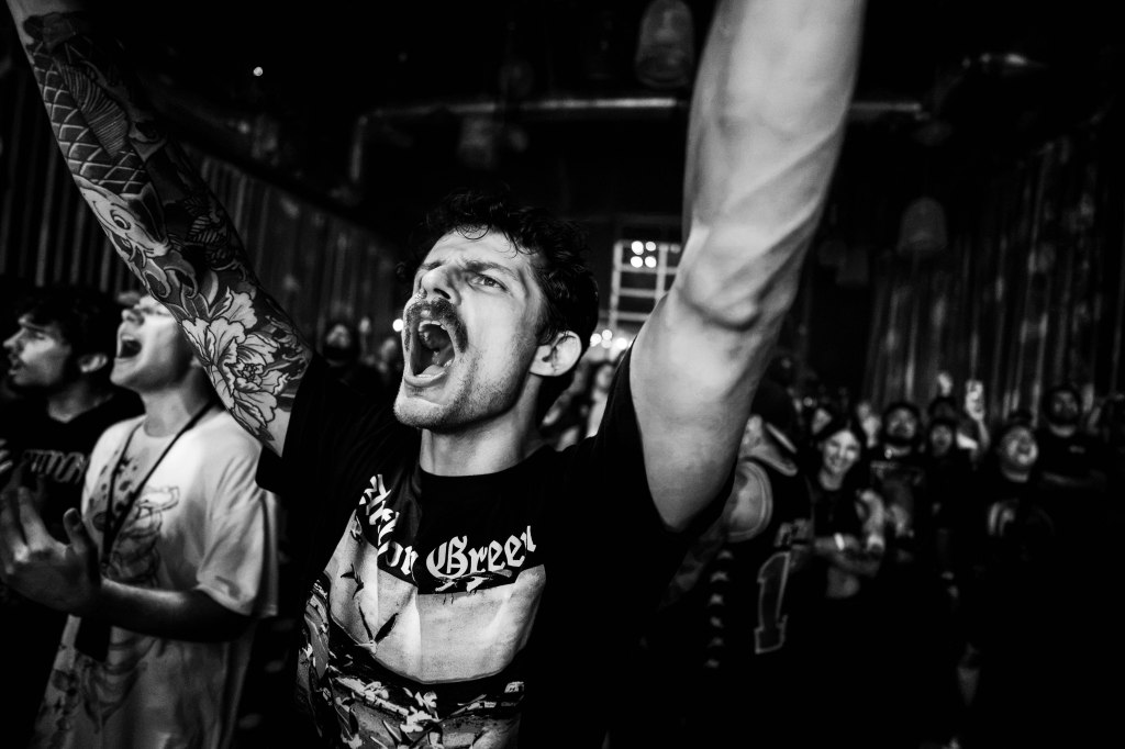 A crowd at a concert with an enthusiastic man in the foreground, arms raised and singing passionately. The image is in black and white, highlighting the excitement of the audience around him.