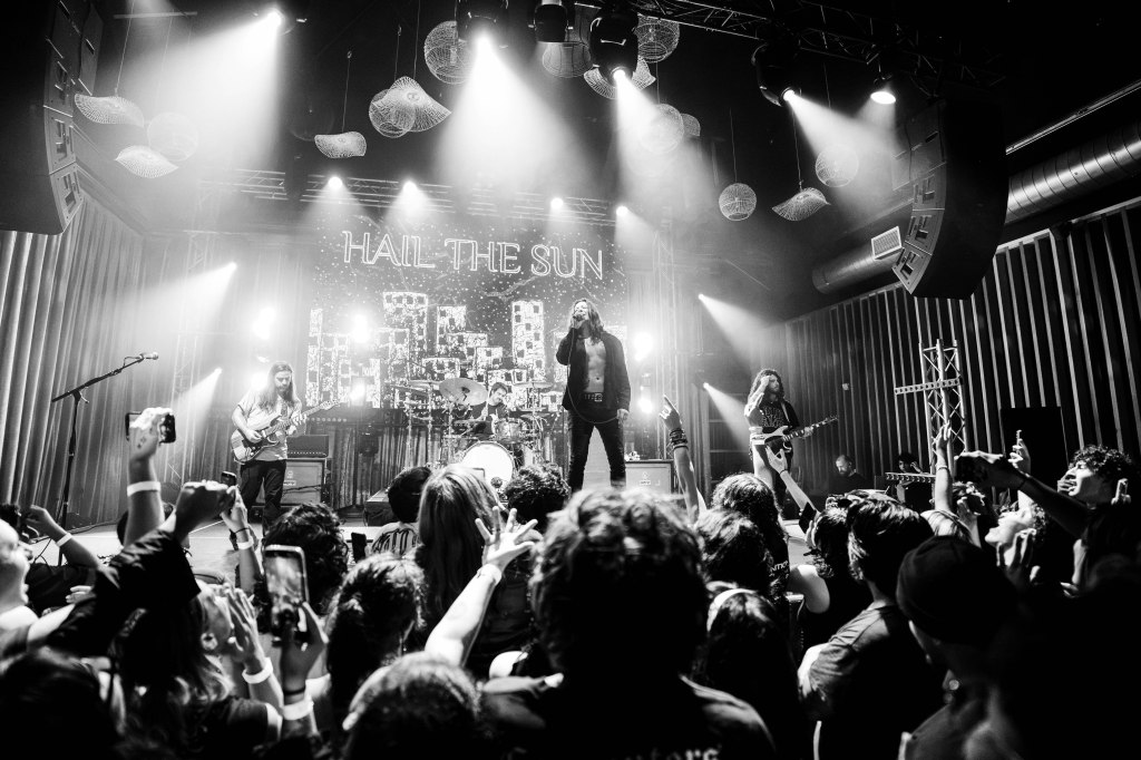 A black and white photo of a live band performing on stage, with a lead singer in front and musicians playing guitars in the background. The audience is engaged, with hands raised and phones capturing the moment. Stage lights illuminate the band against a backdrop featuring the words 'HAIL THE SUN.'