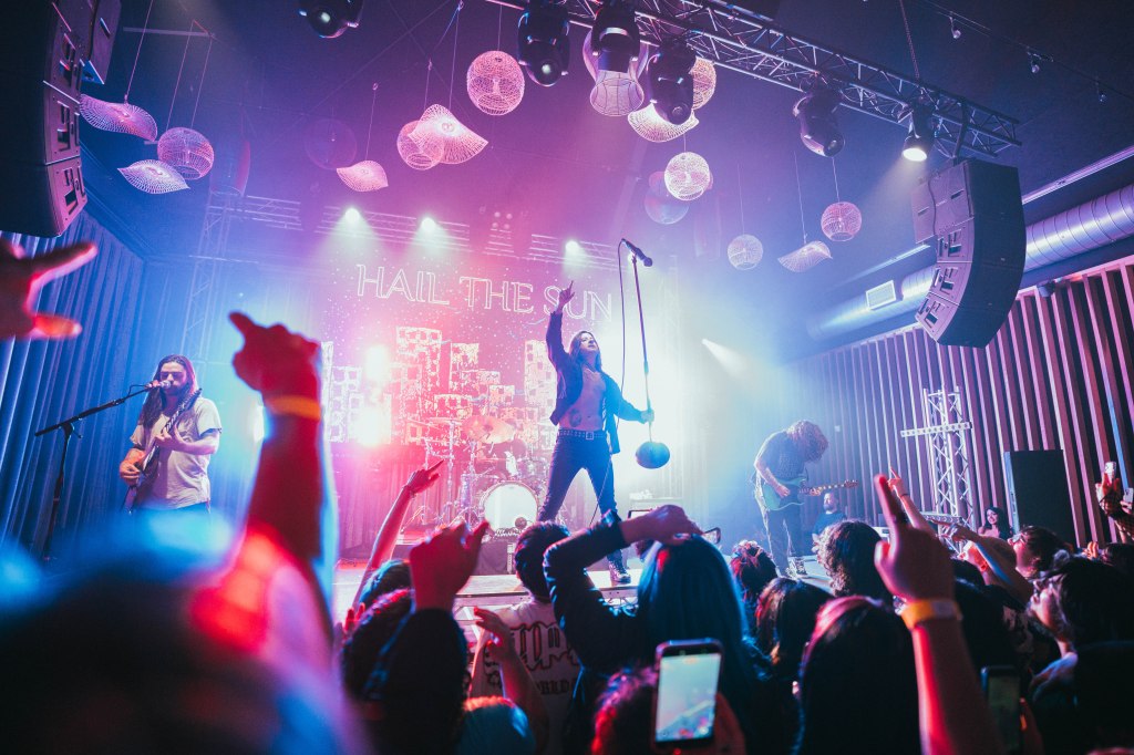A live performance by the band Hail the Sun, featuring a dynamic lead singer on stage, with musicians playing instruments in the background. The audience is engaged, with hands raised and phones in the air, under colorful stage lighting.